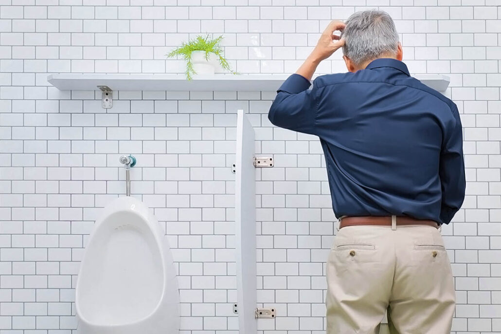 older man with an enlarged prostate stands in front of a urinal waiting to pee