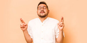 Young man in his 30's with closed eyes and crossed fingers and a hopeful expression