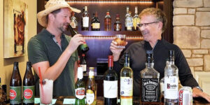 Two men standing at a home bar smile and toast each other—one sipping from a beer bottle and the other holding a short glass with a mixed drink. The counter in front of them displays several alcoholic beverages.