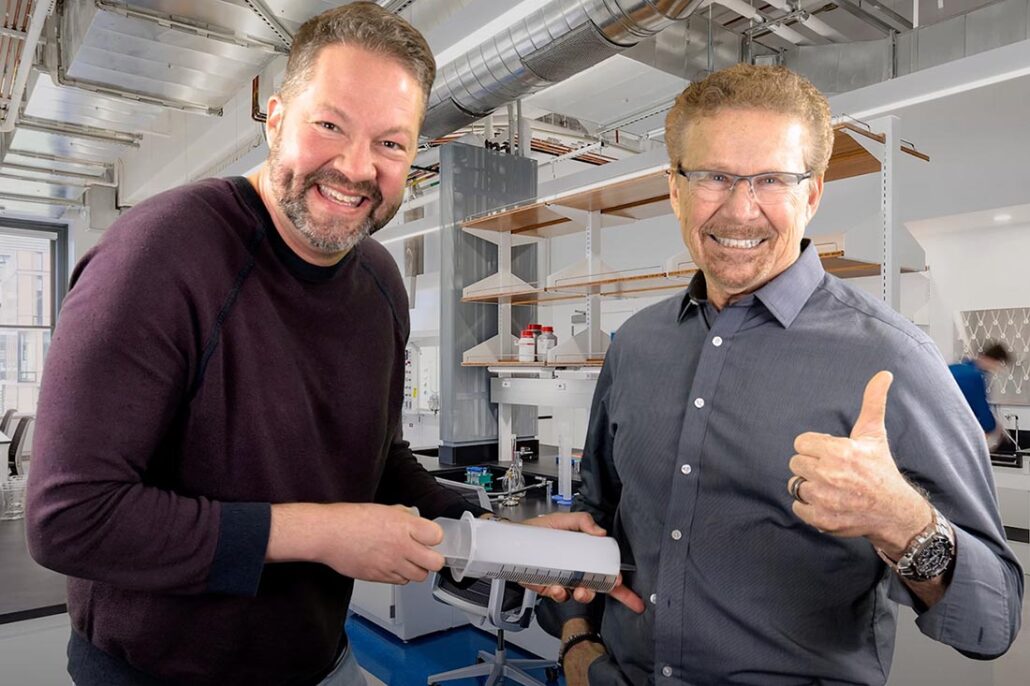 Two men laughing as one inserts an oversized fake syringe into the other's stomach 