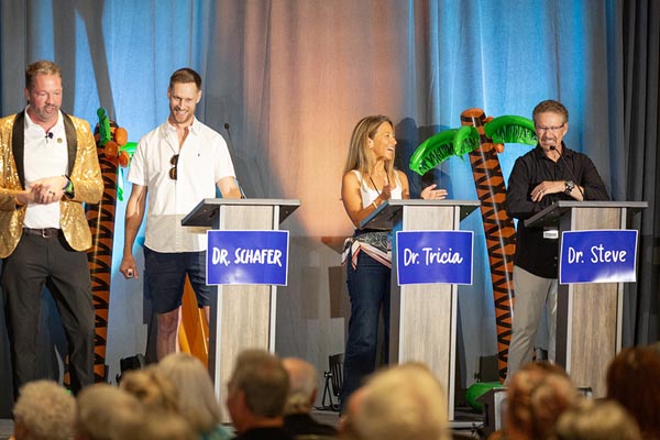 Three contestants and a host on stage stand in front of podiums playing a mock game show