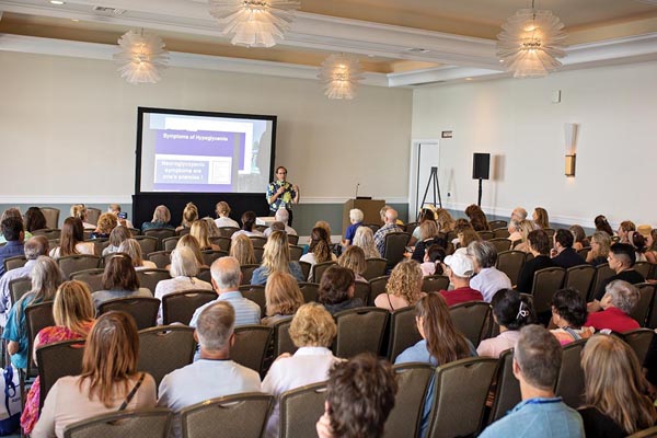 An educatoer stands in front of a large adult crowd giving a talk