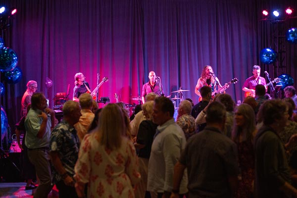 A rock band plays on stage in a banquet room in front of a crowded dance floor
